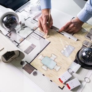 Close-up Of An Architect's Hand Drawing Blueprint With Security Equipment On Desk